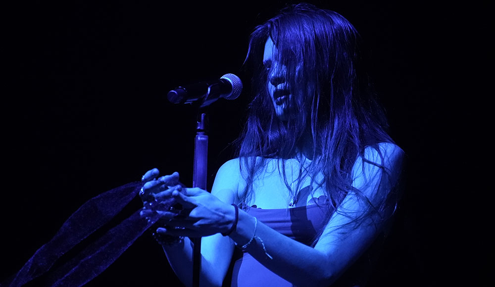 Woman singing into a microphone under a blue spotlight