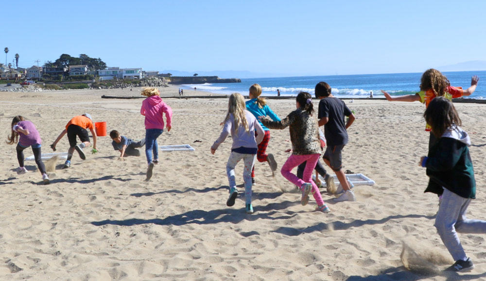 Children of various ages running on the beach and picking up trash