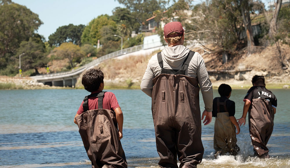 Adult and three kids wearing waders and walking in a river