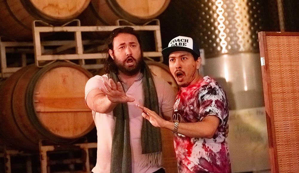 Two men singing in front of a wall of wine barrels