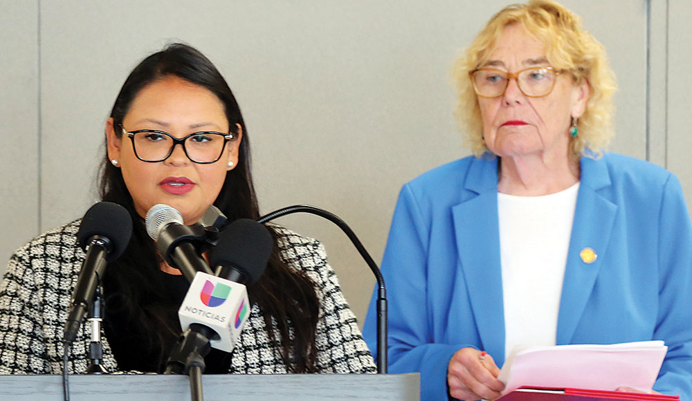 Woman speaking into a microphone standing next to another woman holding a document