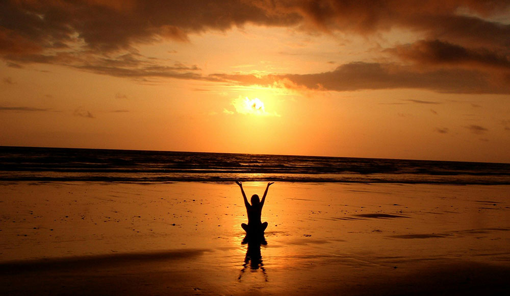 Person sitting on a beach looking at the setting sun, stretching arms skyward