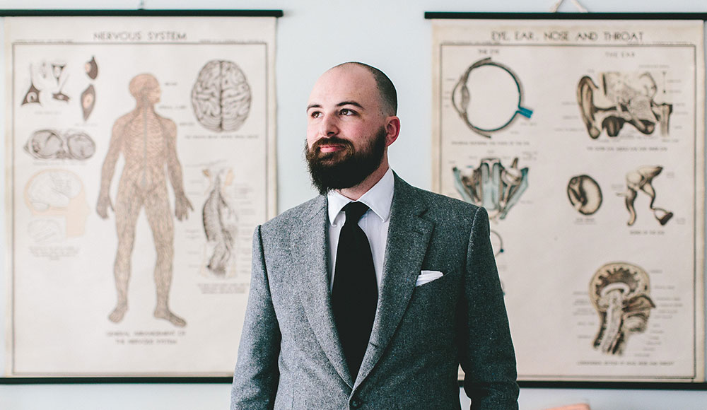 Jesse Thorn in buttoned gray suit, white dress shirt and long black tie stands in front of anatomical posters.