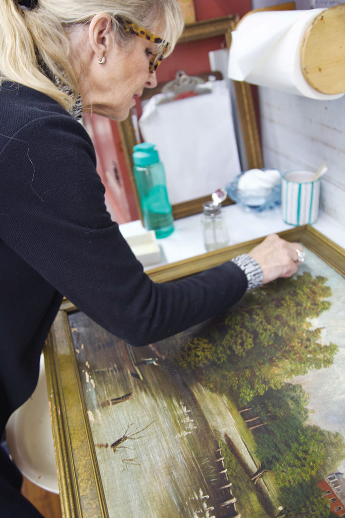 Conservator Diana Wilson carefully restoring an old landscape painting in a frame at an art studio workbench.
