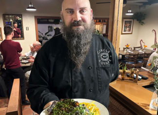 Shaping Up Chef Tyler Reiner holding a plated entrée inside a warmly lit restaurant dining room