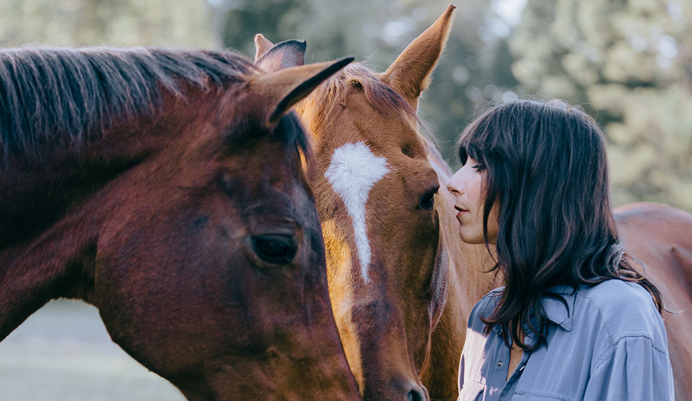 Singer Nicki Bluhm stands closely with two horses, touching foreheads in a peaceful outdoor setting.