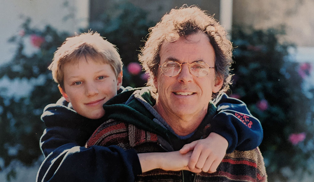 A young Kyle Thiermann hugs his father Eric Thiermann from behind in a sunlit outdoor photo.