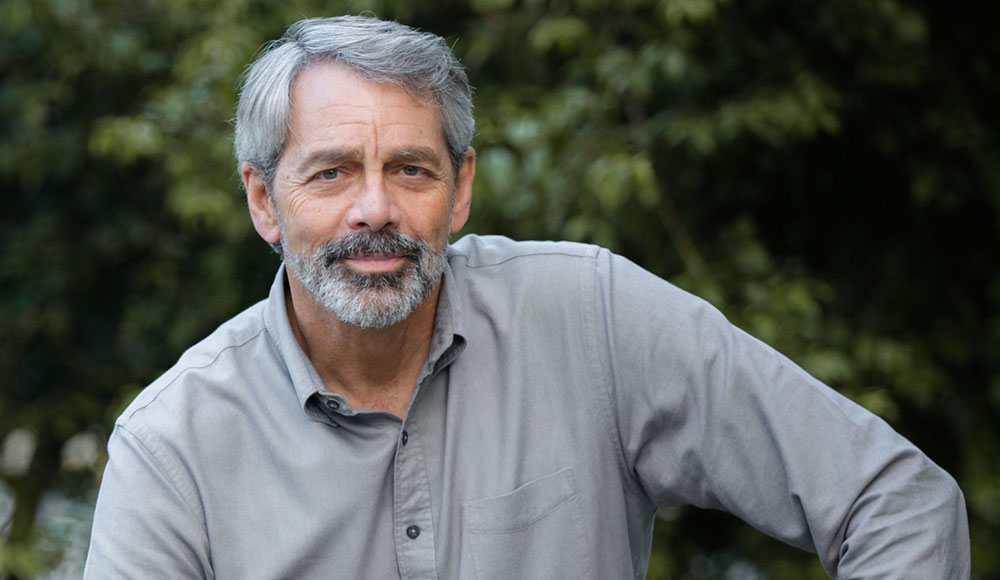 A gray-haired man with a trimmed beard and mustache poses outdoors in a light gray button-down shirt, looking calmly into the camera.