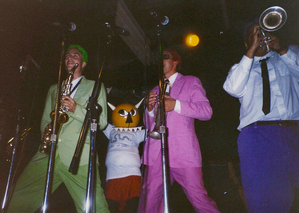 Horn players in brightly colored suits perform onstage beside a costumed character with a large cartoon mask during a Slow Gherkin concert.