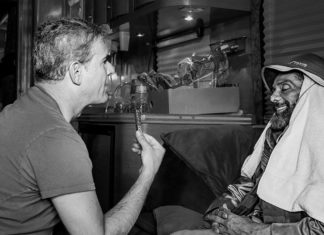 The Editor’s Desk A man sits across from another man wrapped in towels, holding up a harmonica during an interview inside a dimly lit room or trailer.
