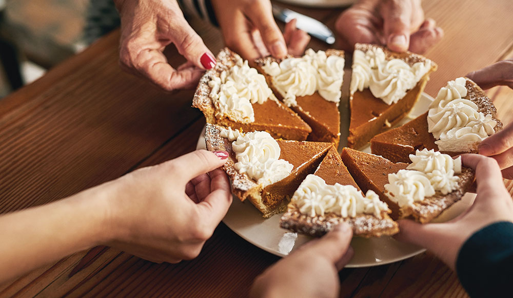 Hands from multiple people reach in to take slices of a pumpkin pie topped with whipped cream at a holiday gathering.